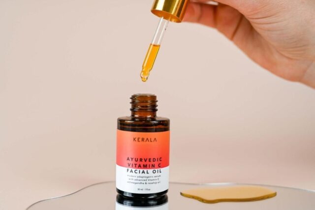 kb-review-hero-1068 A hand holding a glass dropper above an open amber bottle of Kerala Ayurvedic Vitamin C Facial Oil, with oil dripping into a pool on a mirrored surface